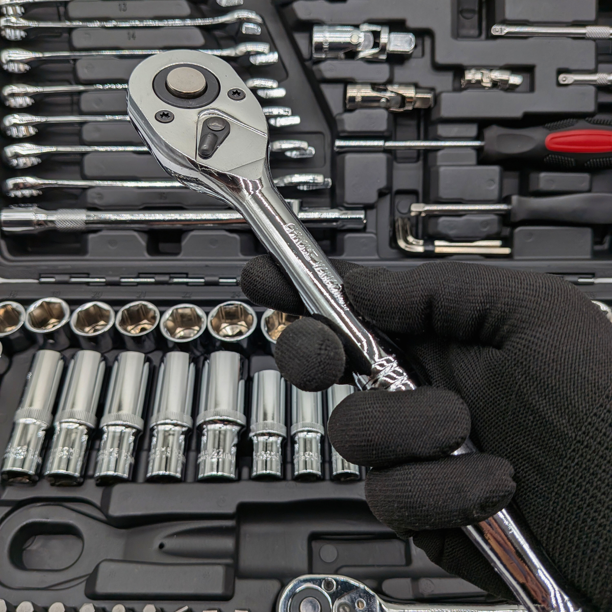 Gloved hand holding a chrome ratchet above the open IRONVOLT 218-piece tool set.